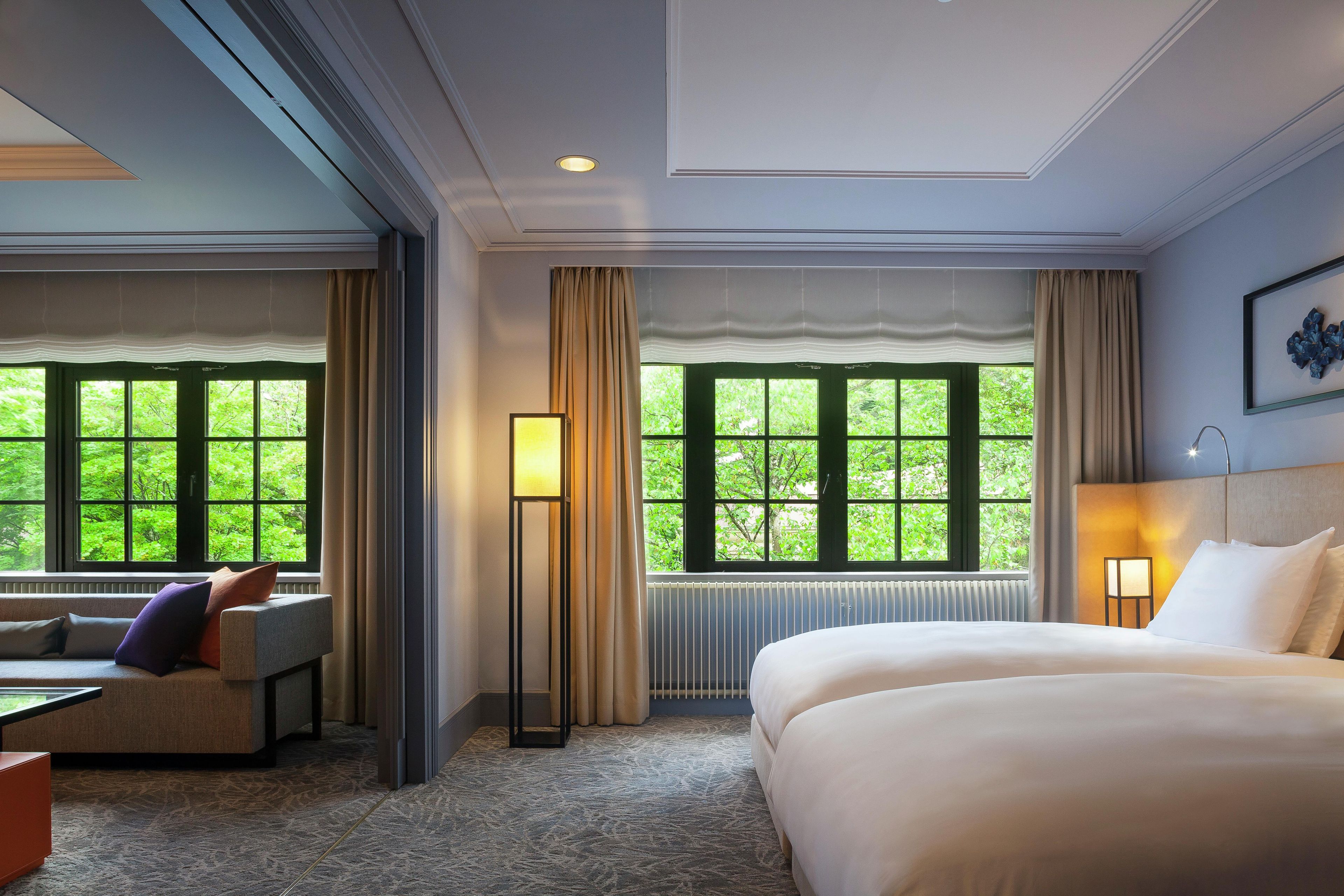 A hotel room with a bed, a desk, a chair, and a view of greenery through large windows.