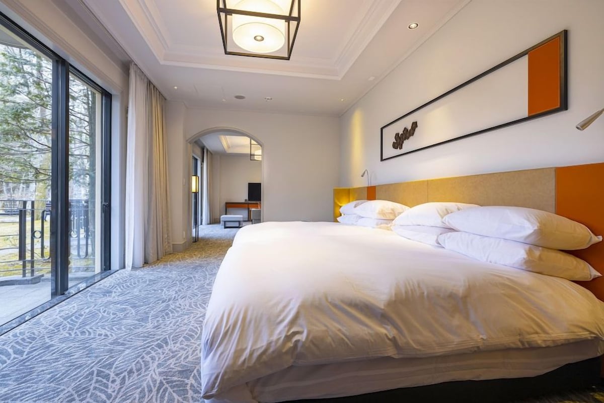 Ein Hotelzimmer mit einem großen Bett, Blick durch das Fenster und einem zur anderen Zimmer führenden Flur.