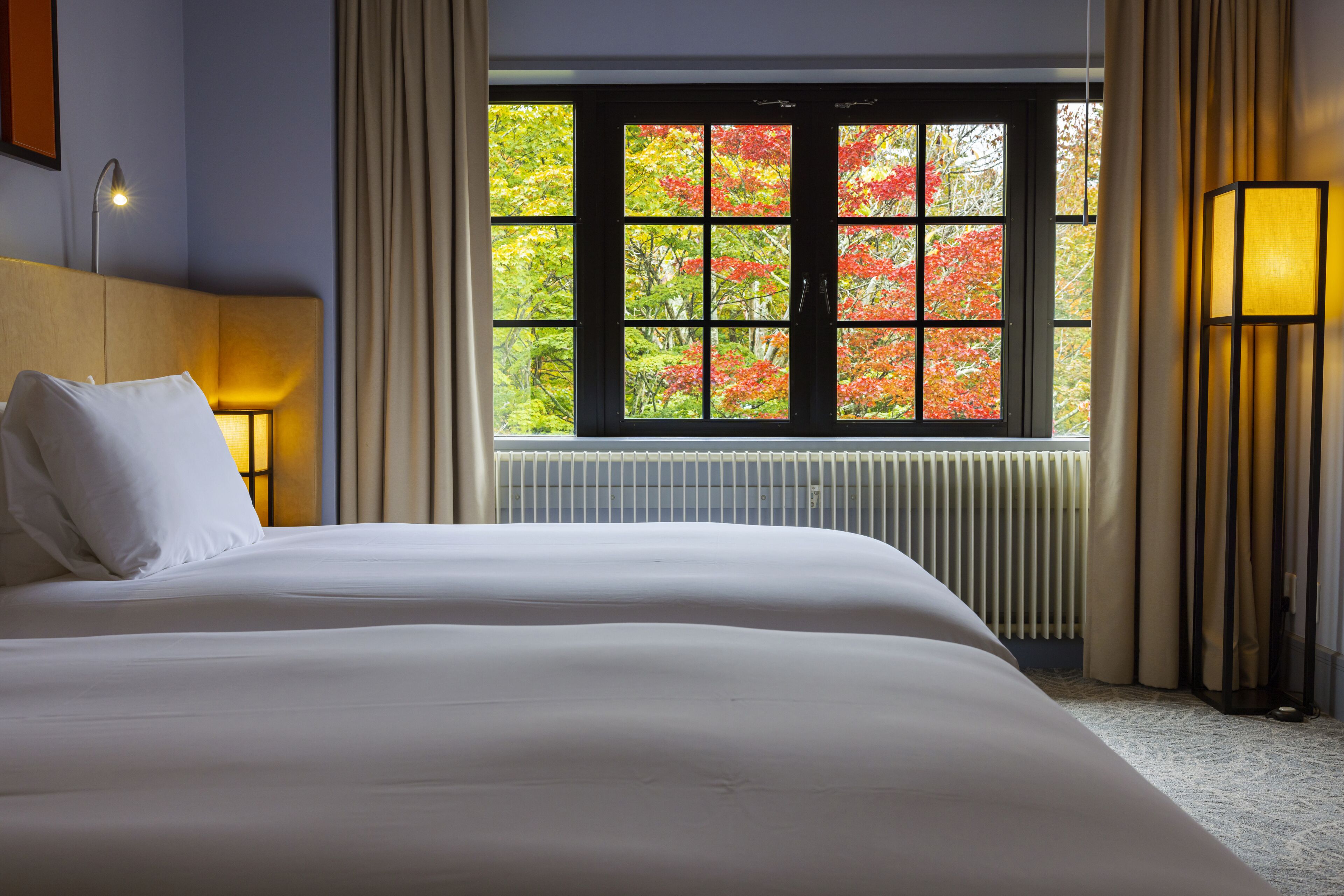 Ein Hotelzimmer mit einem großen Bett, einem Fenster mit Blick auf Herbstlaub, einem Heizkörper und einer Lampe.