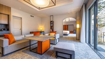 A modern hotel room with a sofa, ottoman, and a small table. There is a bookshelf and a door leading to another room.