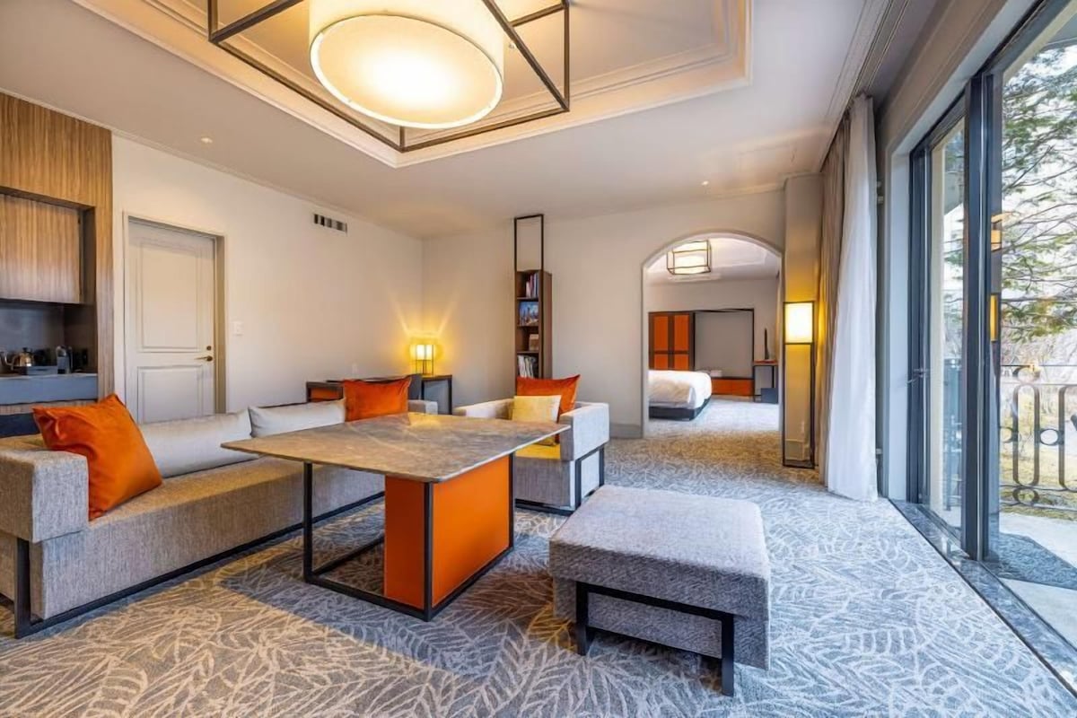 Ein modernes Hotelzimmer mit einer Couch, einem Hocker und einem kleinen Tisch. Ein Bücherregal und eine Tür sind sichtbar.