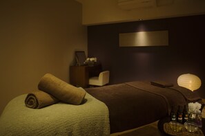 Un cuarto de spa con una camilla de masaje, toallas, una mesita con botellas y una bandeja de aceites, una silla y una lámpara fijada a la pared.