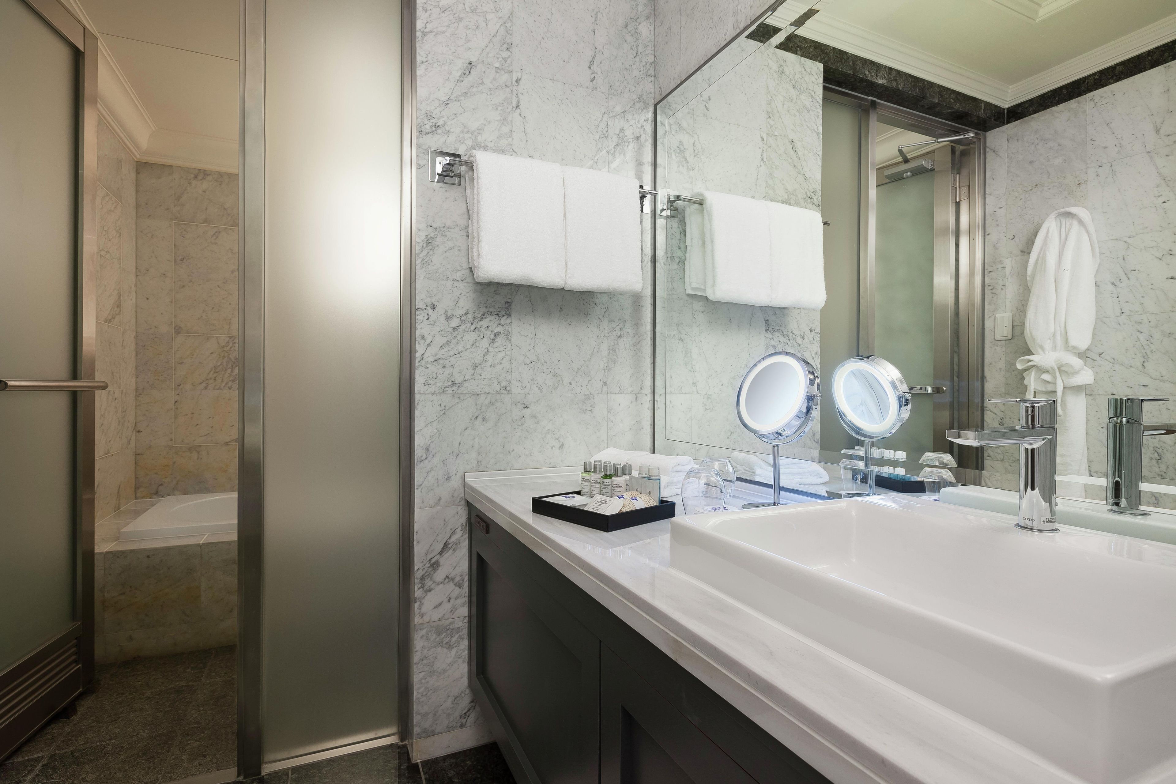 A modern bathroom with a large white sink, two mirrors, a glass-enclosed shower, and marble walls.