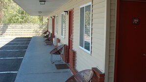 Un motel con una fila de habitaciones, cada una con una puerta roja y una ventana. Hay sillas al aire libre y un paseo cubierto.