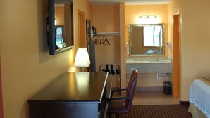 Habitación de hotel con cama, escritorio, silla, lámpara, espejo, lavamanos y toallero.