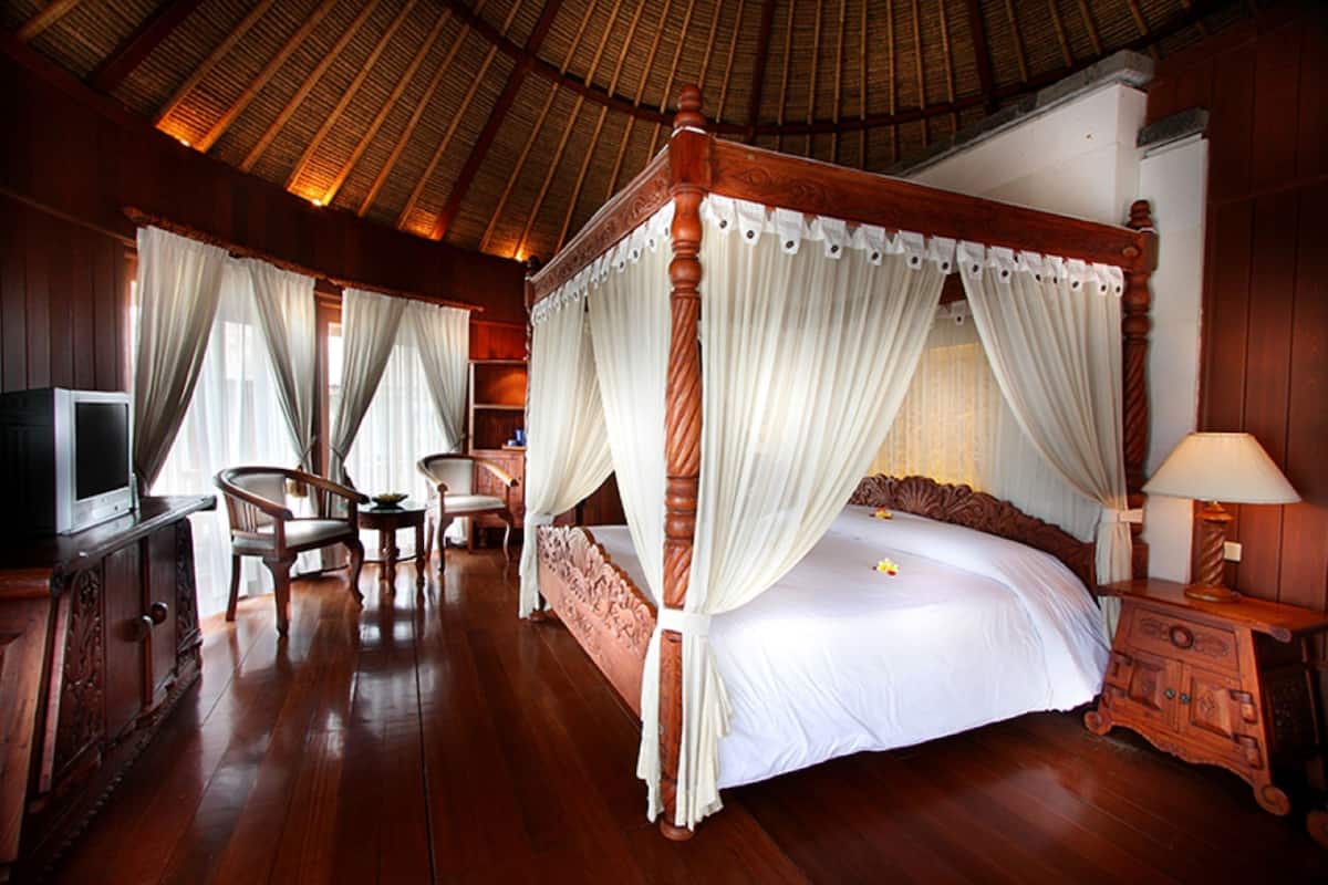 Een bed met hemelbed voorzien van witte gordijnen, een televisie op een houten kast en een nachtkastje met een lamp.