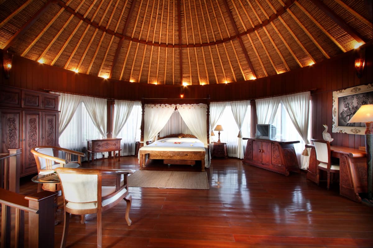Een ruime kamer met een groot bed, houten meubels en een hoog plafond met rieten dak.