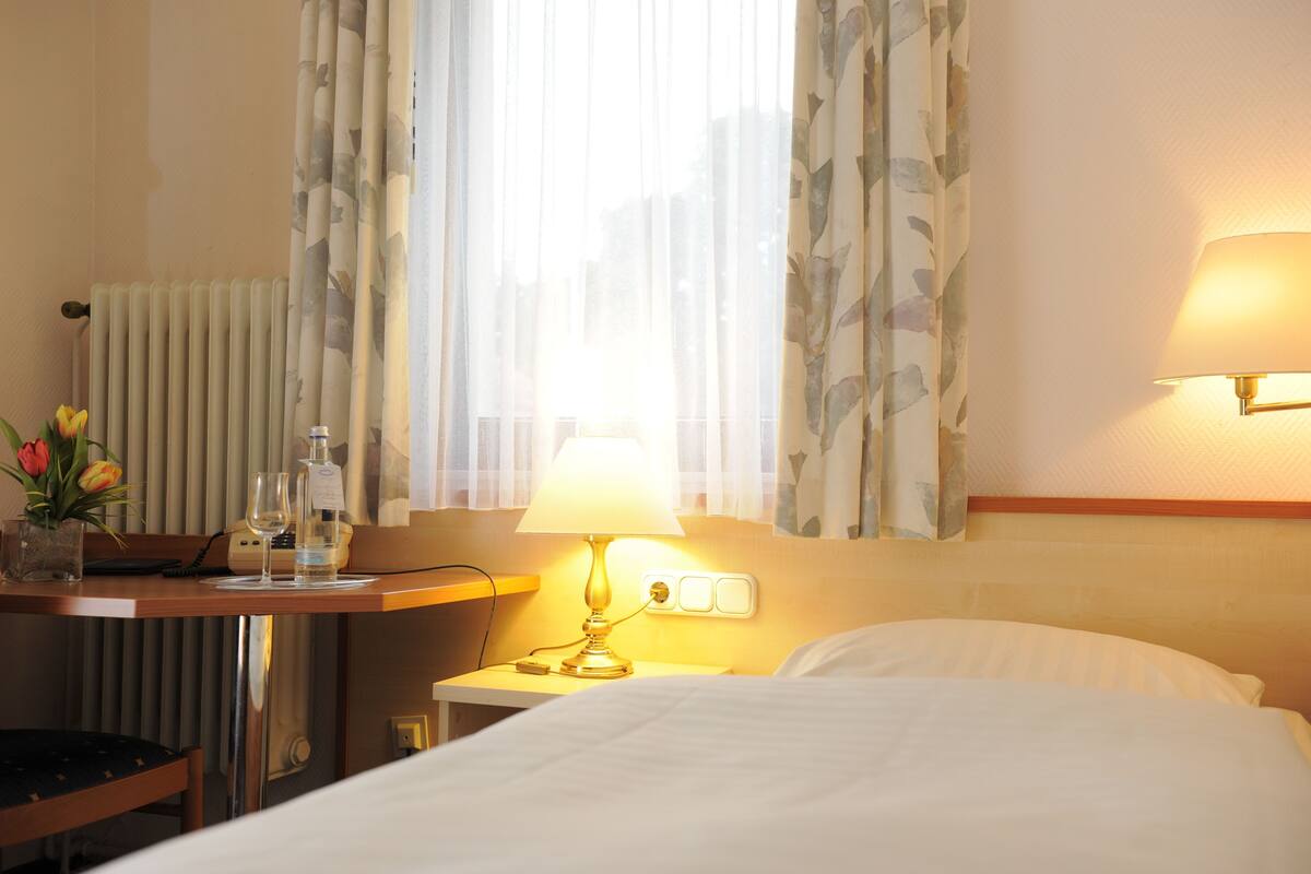 A hotel room with a bed, a bedside table with a lamp, a vase with flowers, a bottle, and a glass.