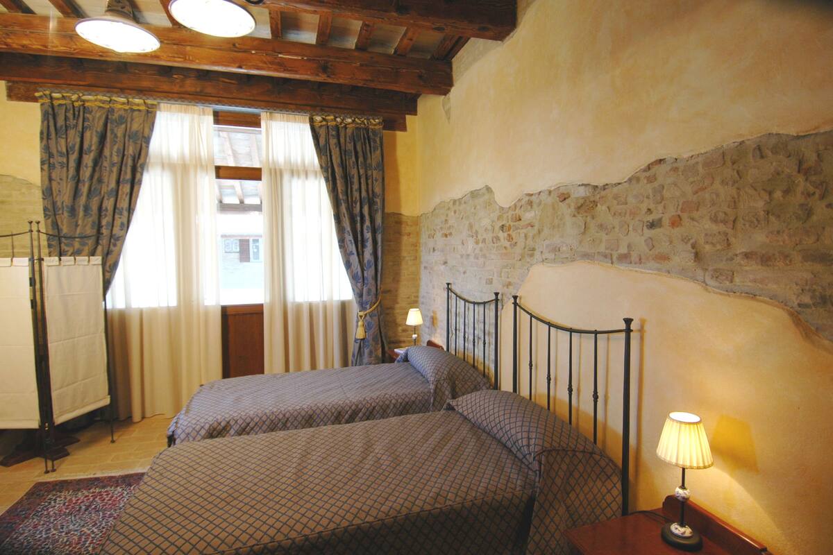 A room with two beds, a wooden ceiling, a stone wall, and a window with curtains.
