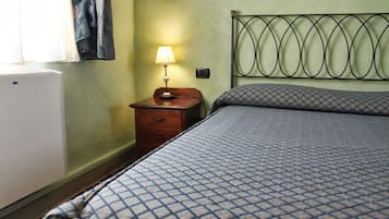 Quarto com cama, mesa de cabeceira com luminária, radiador fixado na parede e janela com cortinas.