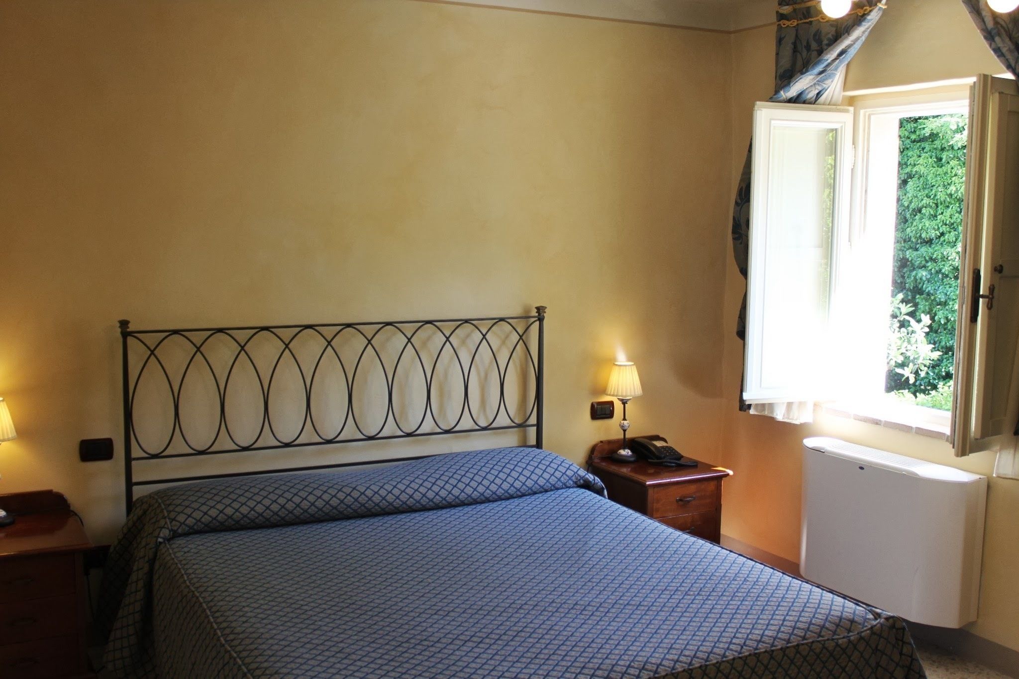 Un dormitorio con cama, lámparas de noche, mesita de noche, ventana con cortinas y un radiador.