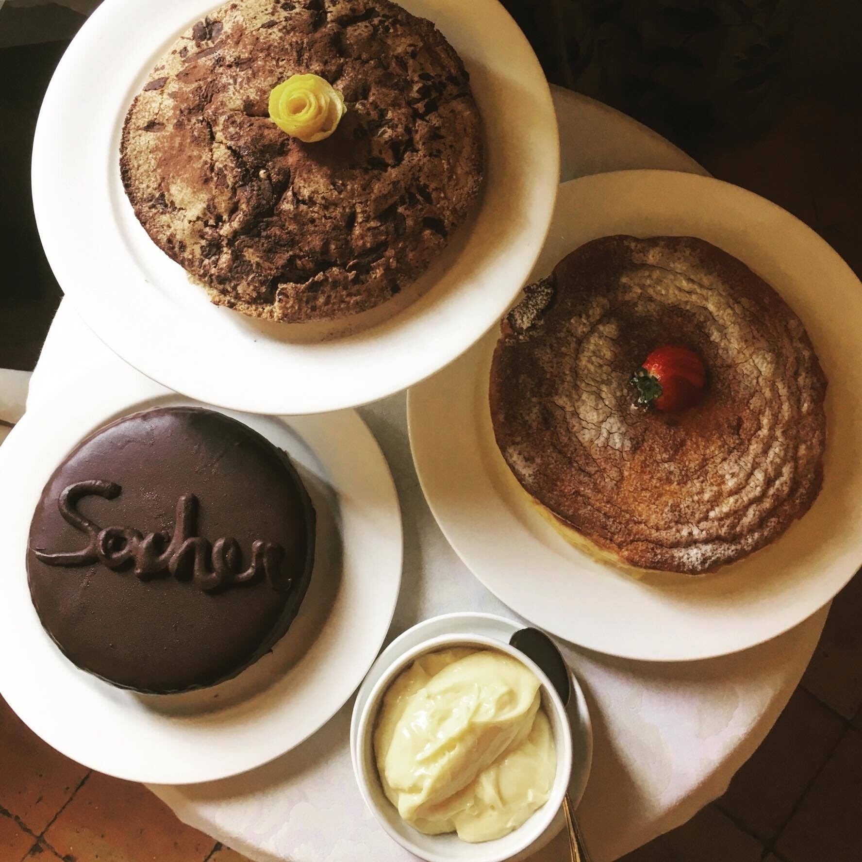 Three cakes on white plates, one with a chocolate surface and the word "Sacher" on it, another with a cherry on top, and a third with a yellow flower decoration.