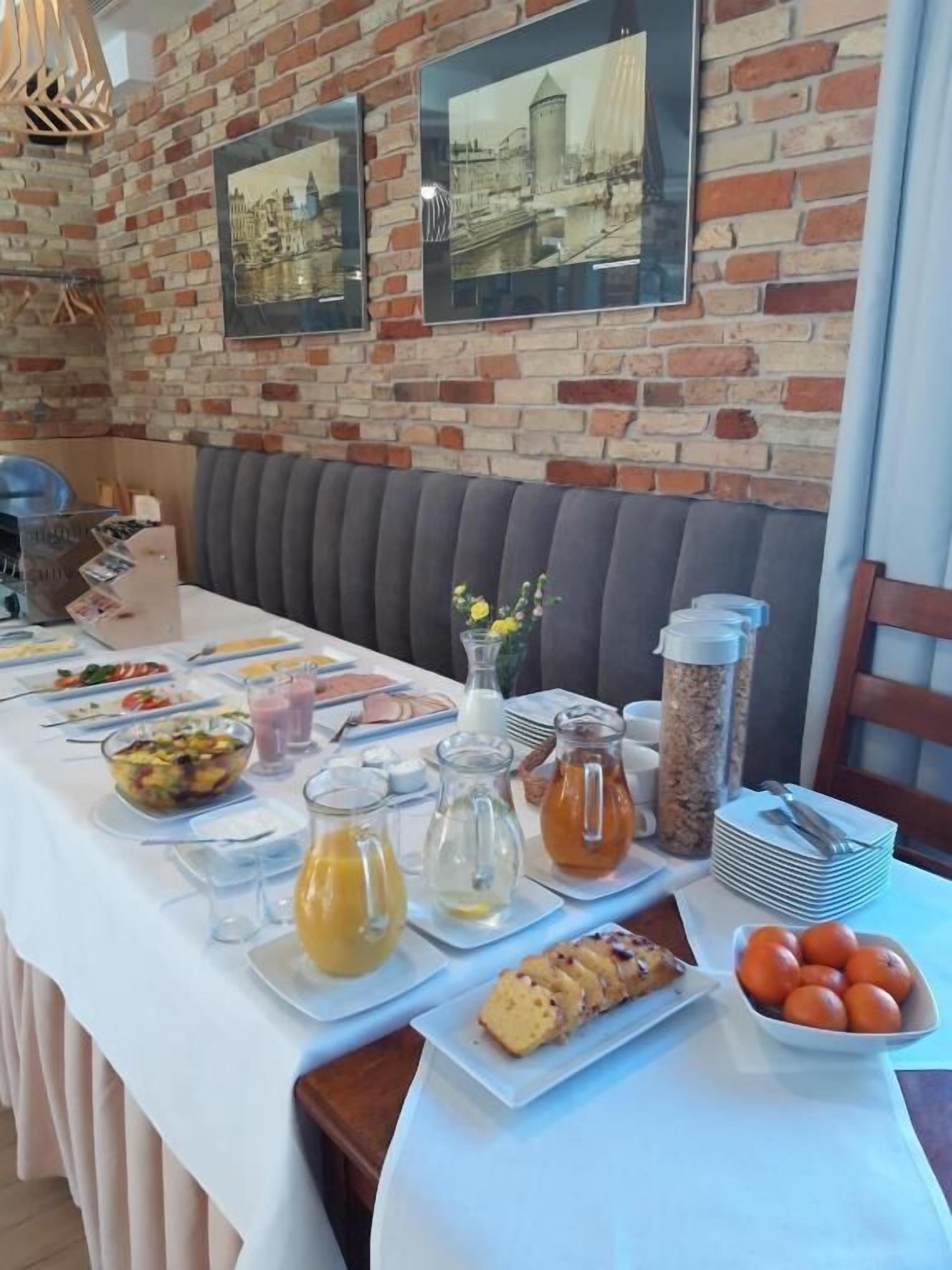 Buffet de café da manhã com diversos alimentos e bebidas, em frente a uma parede de tijolos com quadros.