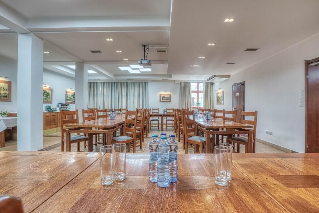 A spacious dining area with wooden tables and chairs, a projector on the ceiling, and a view of the kitchen in the background.