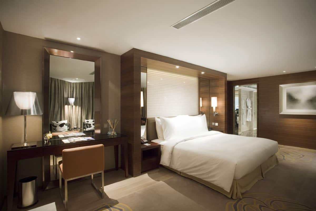 Habitació d'hotel moderna amb un llit gran, un escriptori amb una cadira, un tocador amb un mirall i una finestra ampla amb persianes.