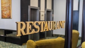 Aula, jossa on kuvioitu matto ja keltainen nojatuoli, ja seinällä on kyltti, jossa lukee: ”RAVINTOLA”.