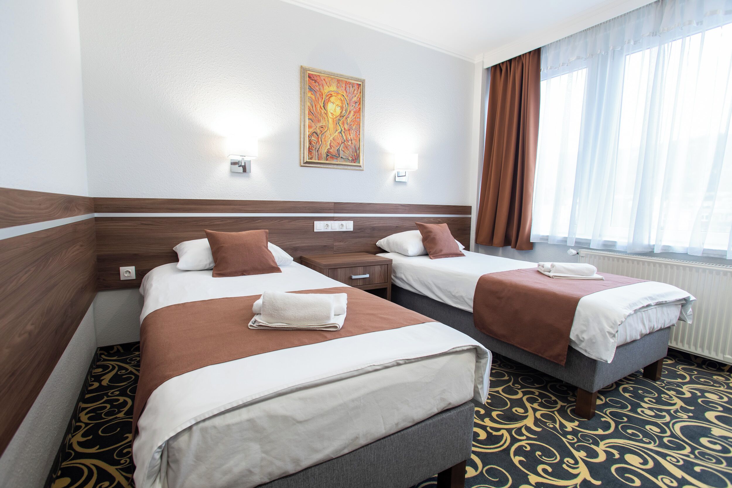 A hotel room with two beds, a painting on the wall, and a window with curtains.