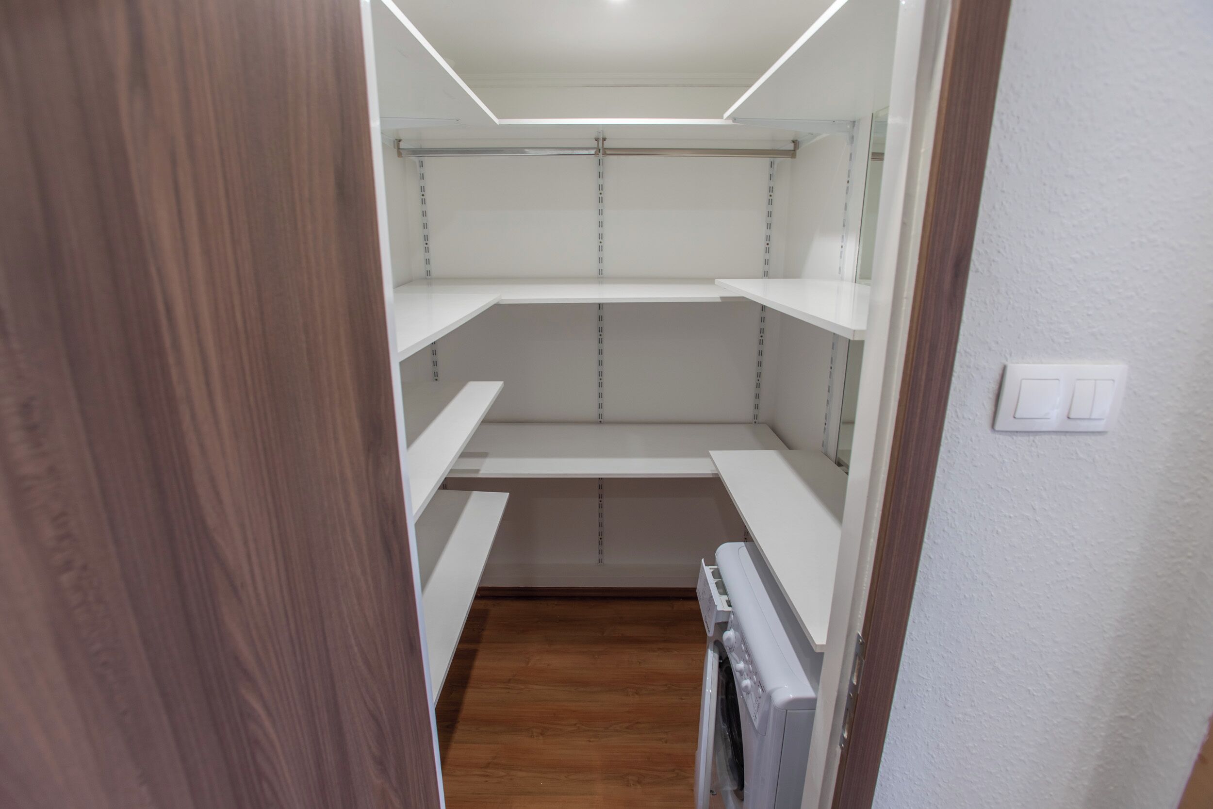 A narrow hallway with wooden flooring, white walls, and a white ceiling with recessed lighting.