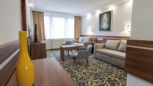 Hotellihuone, jossa on puinen ruokapöytä, sohva, pieni pöytä tuoleineen ja suuri ikkuna, jossa on verhot.