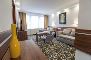 Quarto de hotel com mesa de jantar de madeira, sofá, mesa pequena com cadeiras e uma janela grande com cortinas.