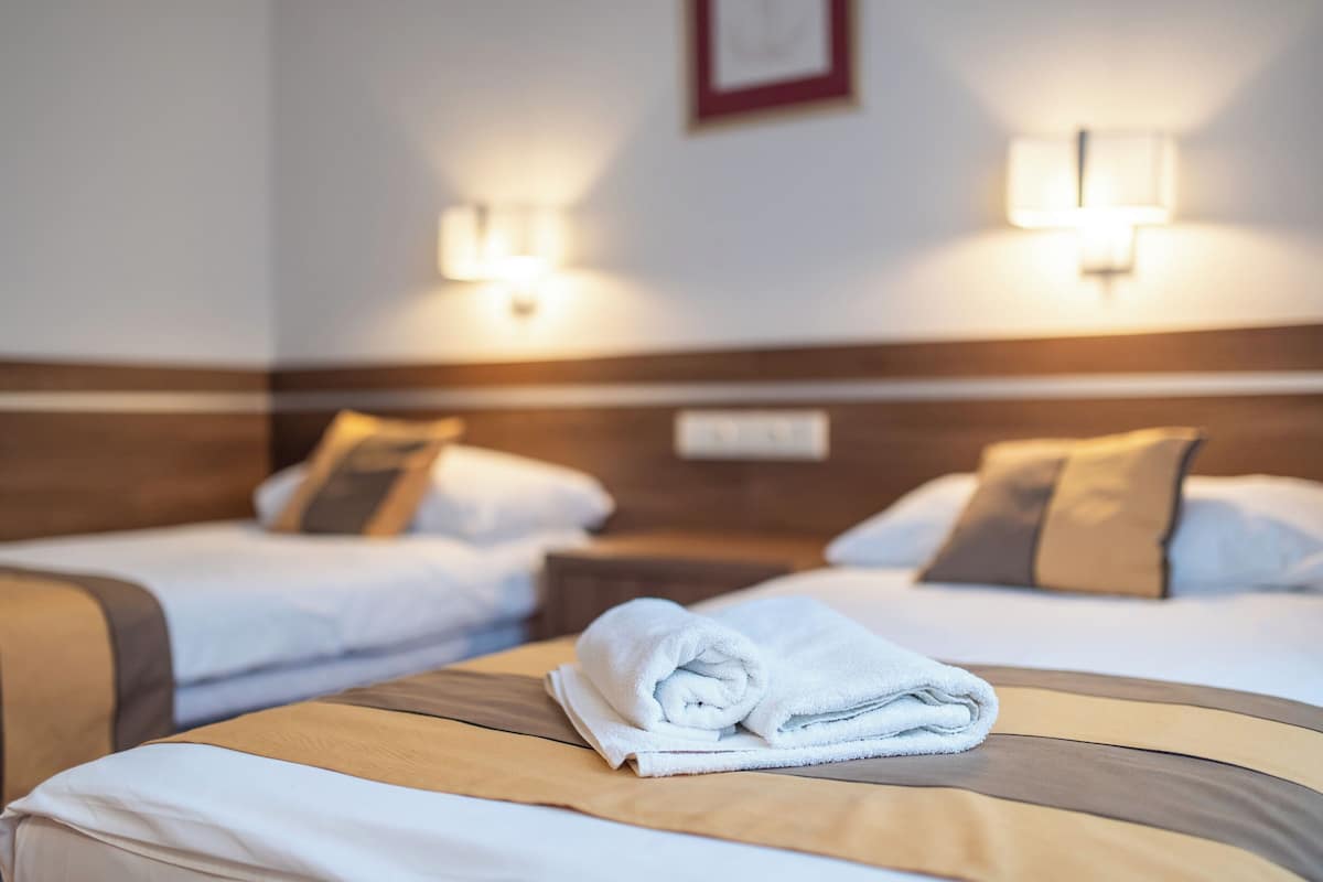 Ett hotellrum med två sängar, var och en med en sänggavel, vita sängkläder och en hoprullad handduk på sängen. På vardera sidan av sängarna finns väggmonterade lampor.