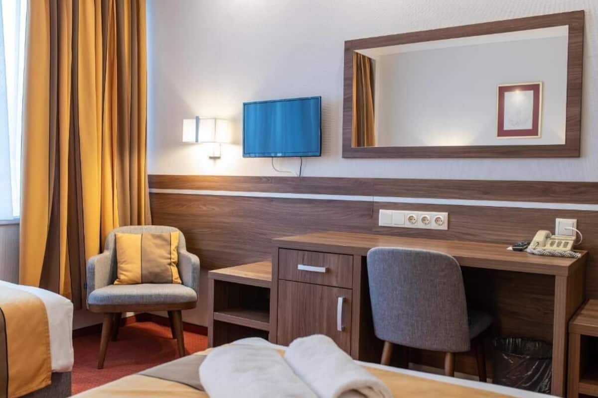 Ett hotellrum med en säng, ett skrivbord med en stol, en tv, en spegel och en telefon.