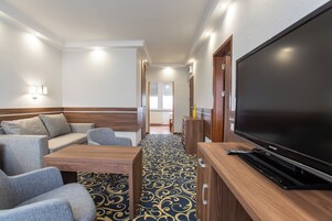 Quarto de hotel com uma televisão de tela grande, um sofá, uma mesa de centro e um tapete estampado.