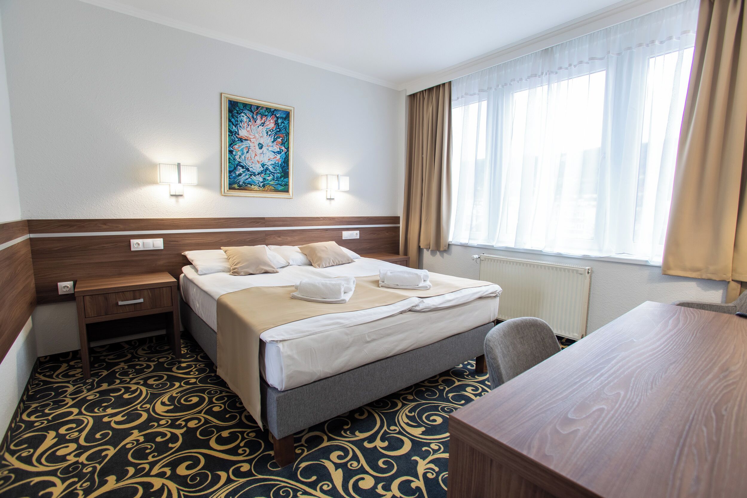 Ein Hotelzimmer mit Bett, Nachttischen, Schreibtisch, Stuhl, einem Fenster mit Vorhängen und einem Wandbild.