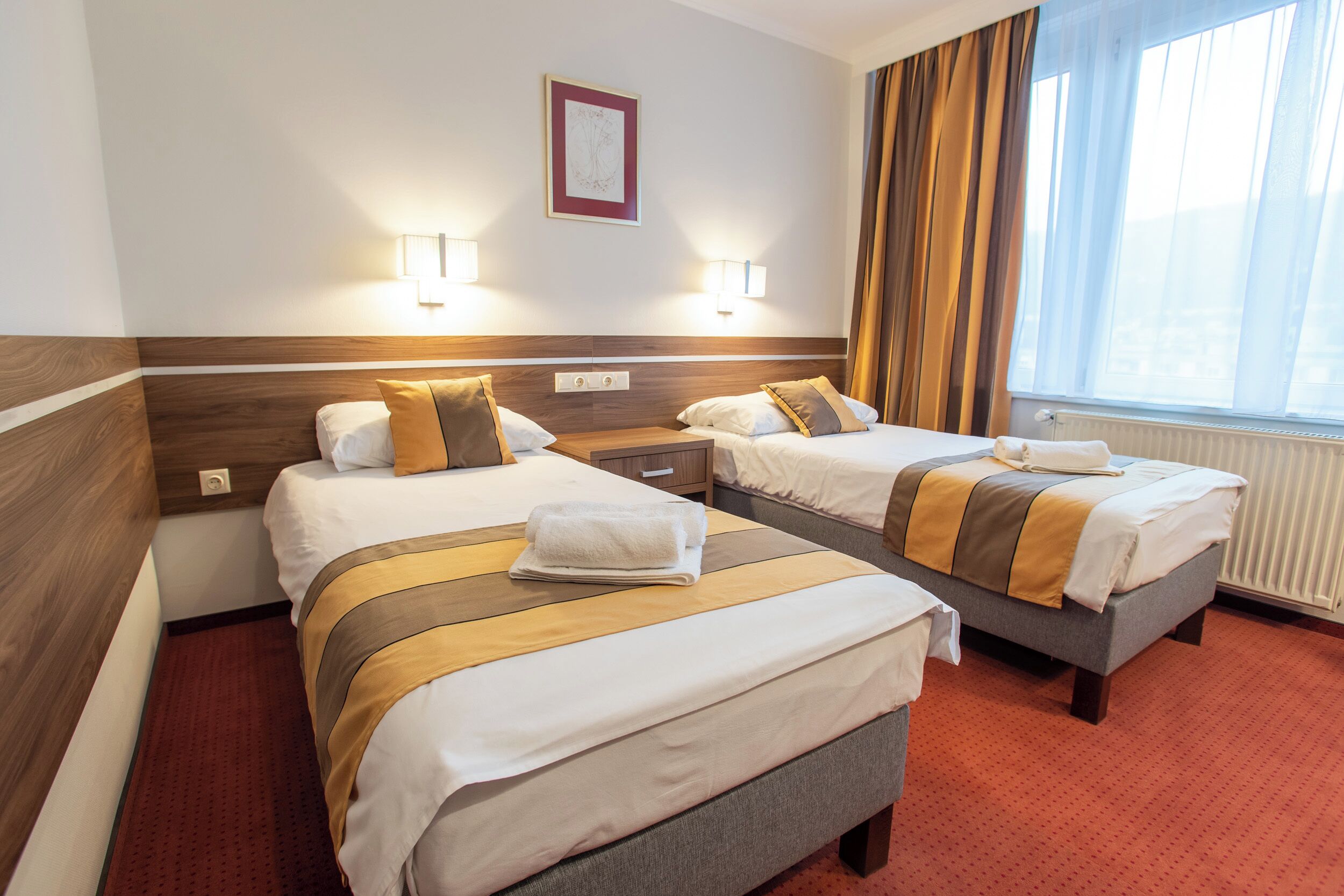 A hotel room with two beds, each with a wooden headboard, a nightstand, two wall-mounted lamps, and a window with curtains.