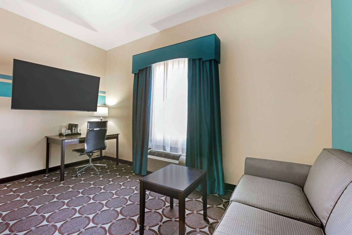 A hotel room with a flat-screen TV, a desk with a chair, a small table, a sofa, and a patterned carpet.