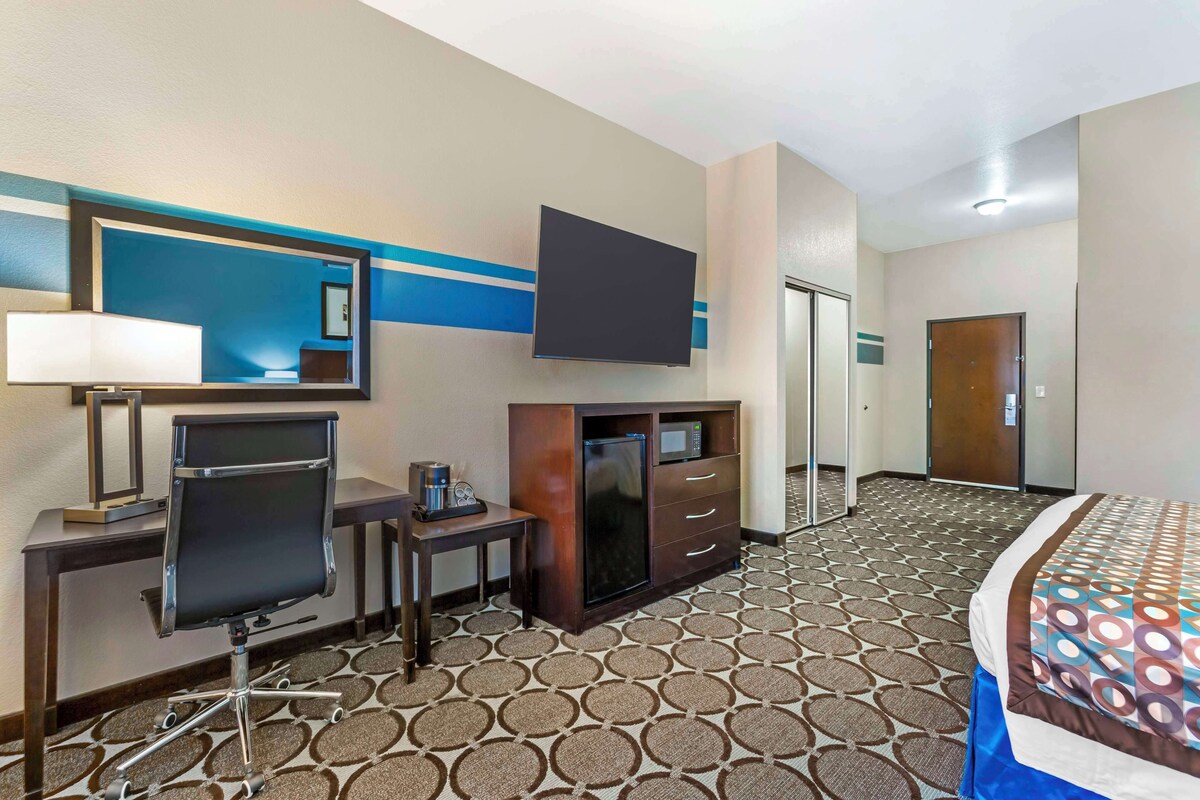 A hotel room with a bed, a desk with a computer, a TV, a minibar, and a door.