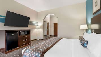 A hotel room with a bed, a flat-screen TV, a minibar, a desk with a lamp, and a patterned carpet.
