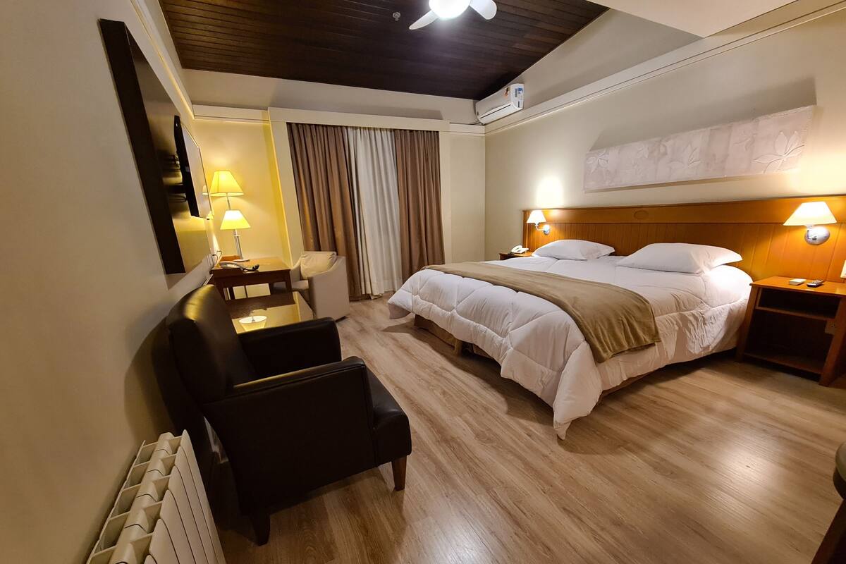 A hotel room with a bed, bedside tables, a desk, a chair, and a radiator.