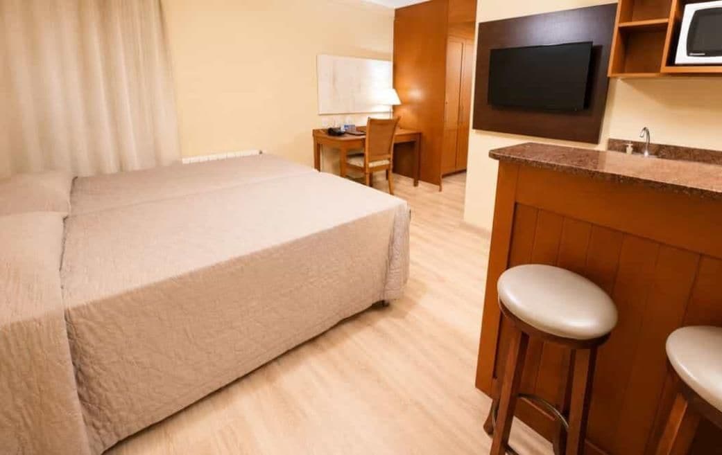 A hotel room with a bed, a desk, a chair, a TV, and a kitchenette with stools.