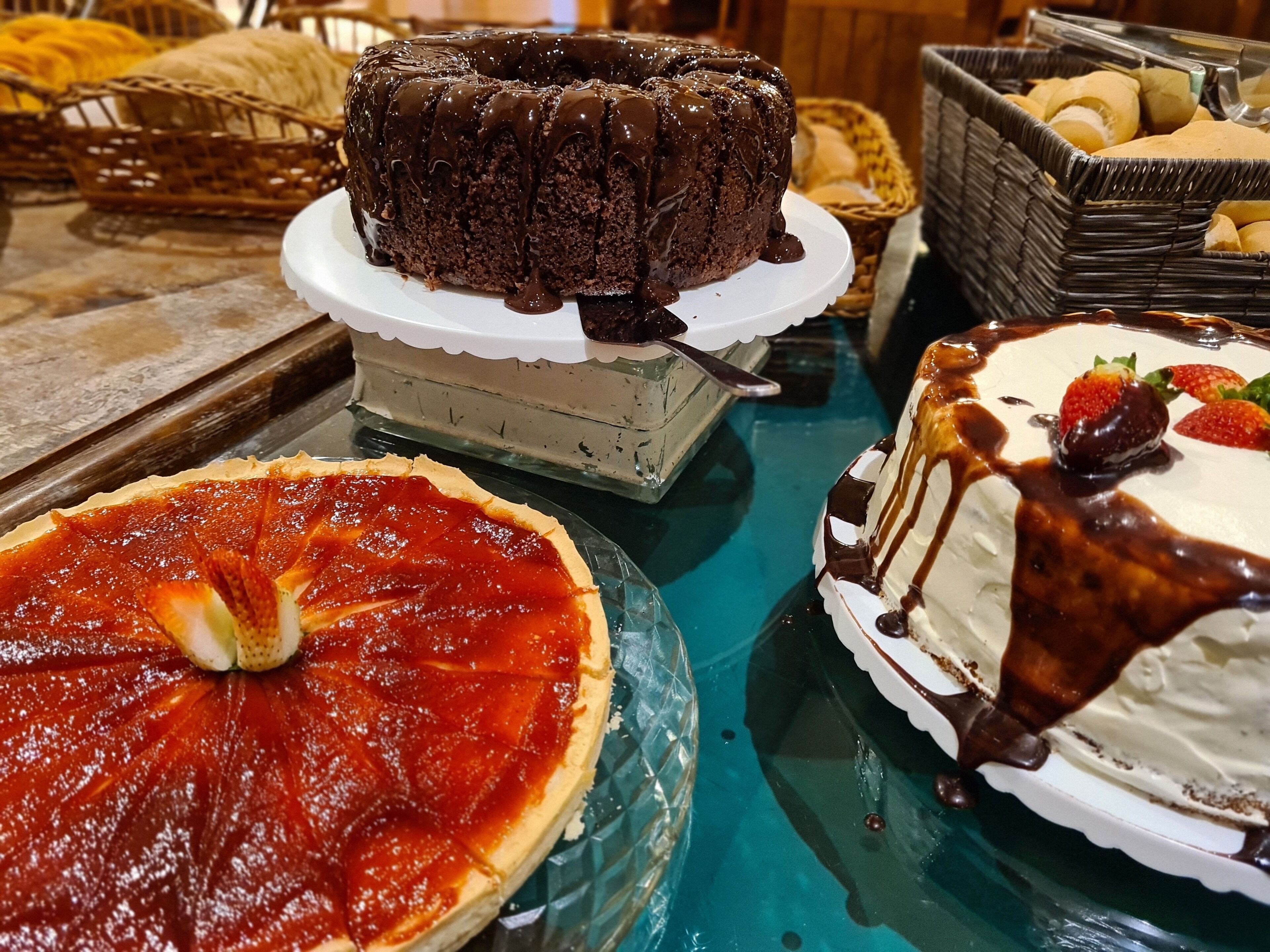 Diversos doces em exposição, incluindo um bolo de chocolate, uma tarte de morango e um bolo branco com calda de chocolate e morangos.