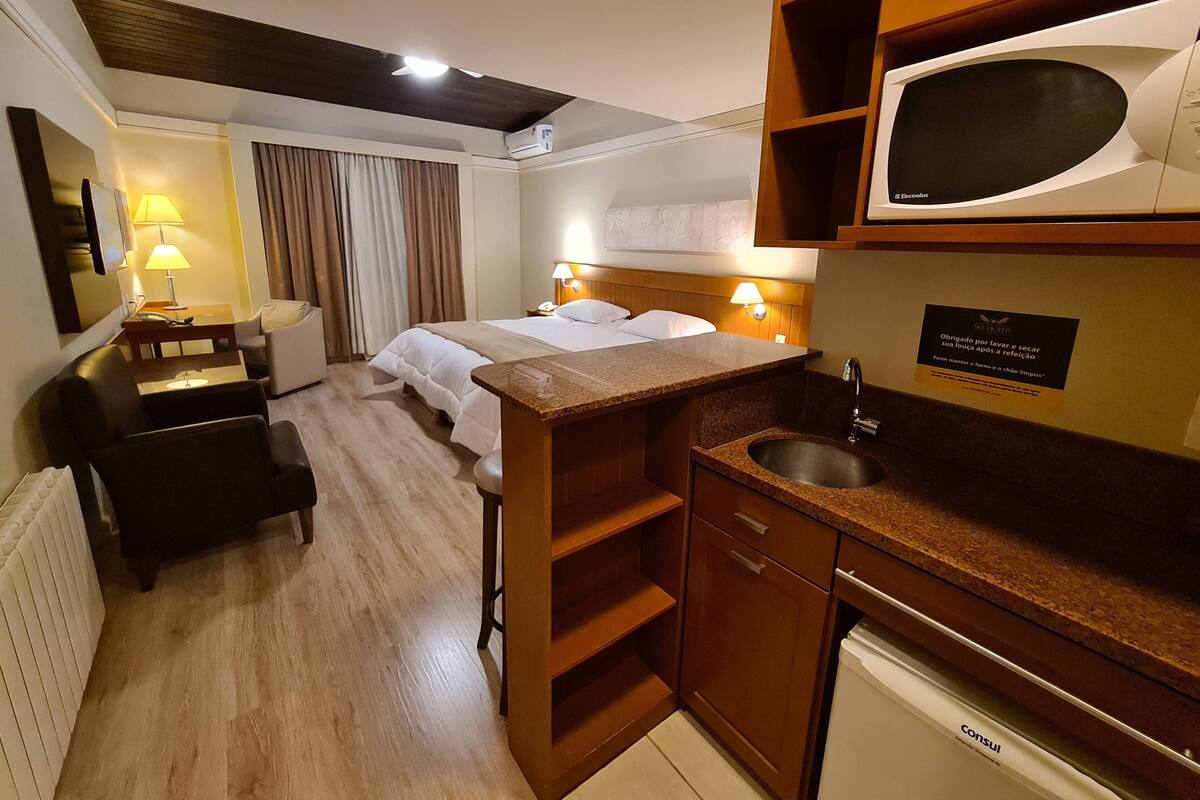 A hotel room with a bed, bedside tables, a desk, a chair, a kitchenette, and a bathroom.