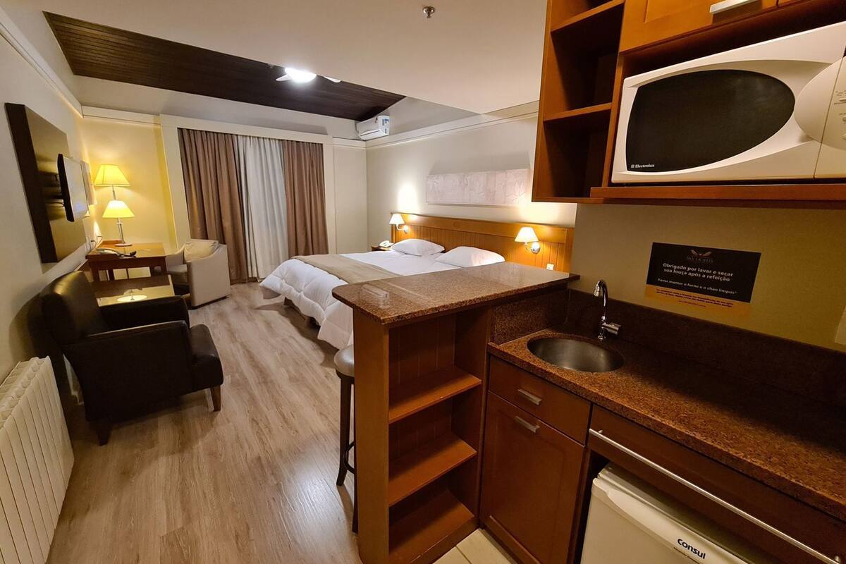 A hotel room with a bed, bedside tables, a desk, a chair, a kitchenette, and a bathroom.