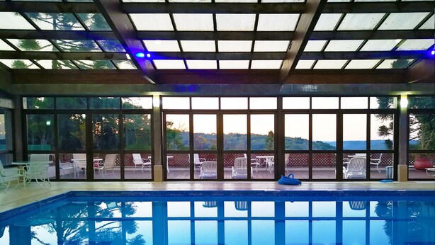 Una piscina interior con paredes de vidrio transparente, vistas al exterior y una zona de terraza con sillas y mesas.