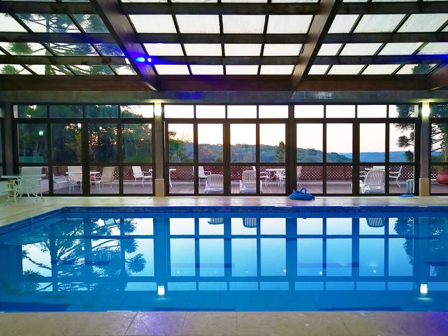Una piscina interior con paredes de vidrio transparente, vistas al exterior y una zona de terraza con sillas y mesas.