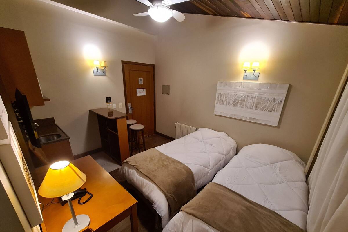 A small hotel room with two beds, a desk with a lamp, a radiator, and a ceiling fan.