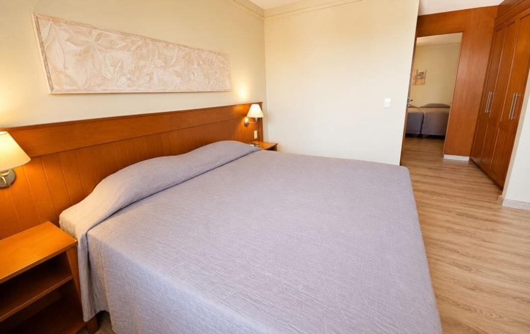 Una habitación de hotel con cama, mesitas de noche, lámpara de pared y armario de madera.