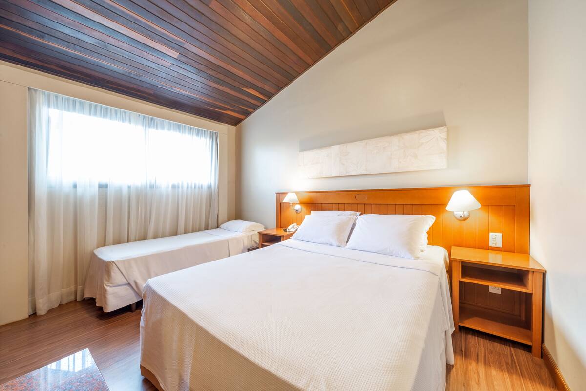 A hotel room with a large bed, a smaller bed, wooden ceiling, and a window with curtains.