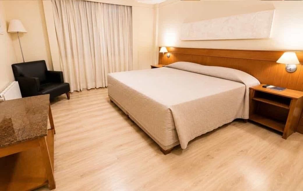 A hotel room with a bed, a chair, a desk, a lamp, and a nightstand.