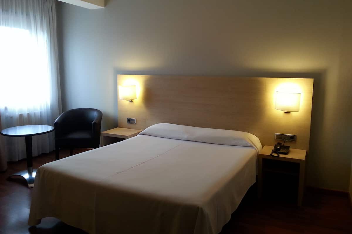 A hotel room with a large bed, two bedside lamps, a chair, a small table, and a window with curtains.