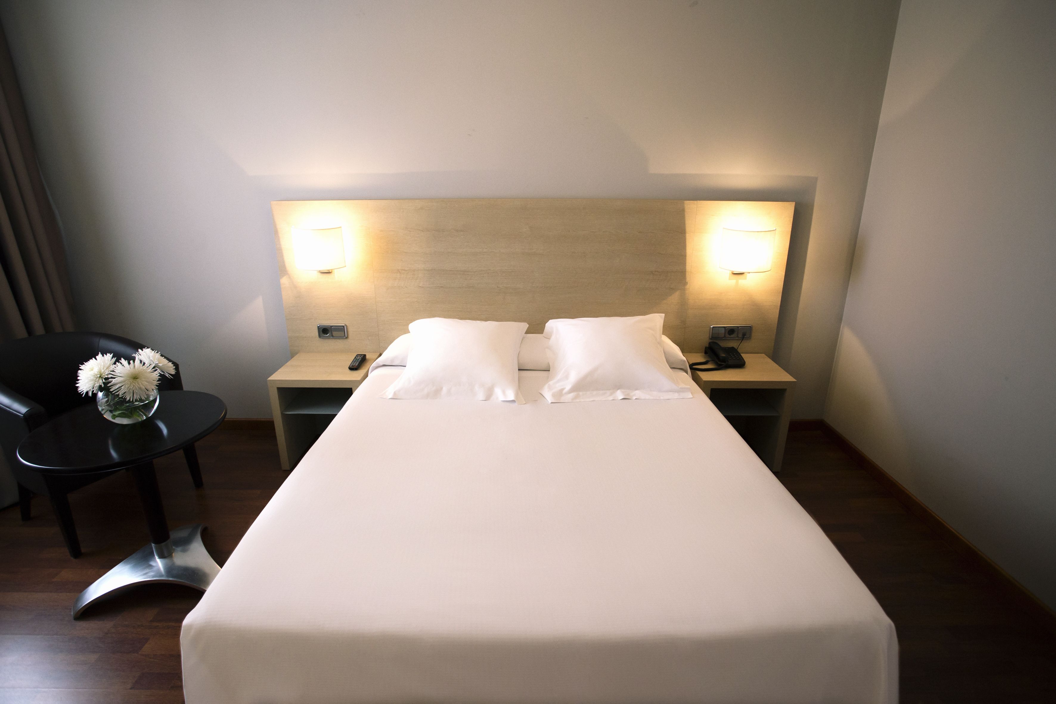 Una habitación de hotel con una cama individual, dos lámparas de noche, una mesita con un jarrón de flores y una silla.