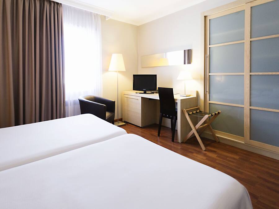 Habitació d'hotel amb dos llits, escriptori amb un ordinador, una cadira i una finestra gran amb cortines.
