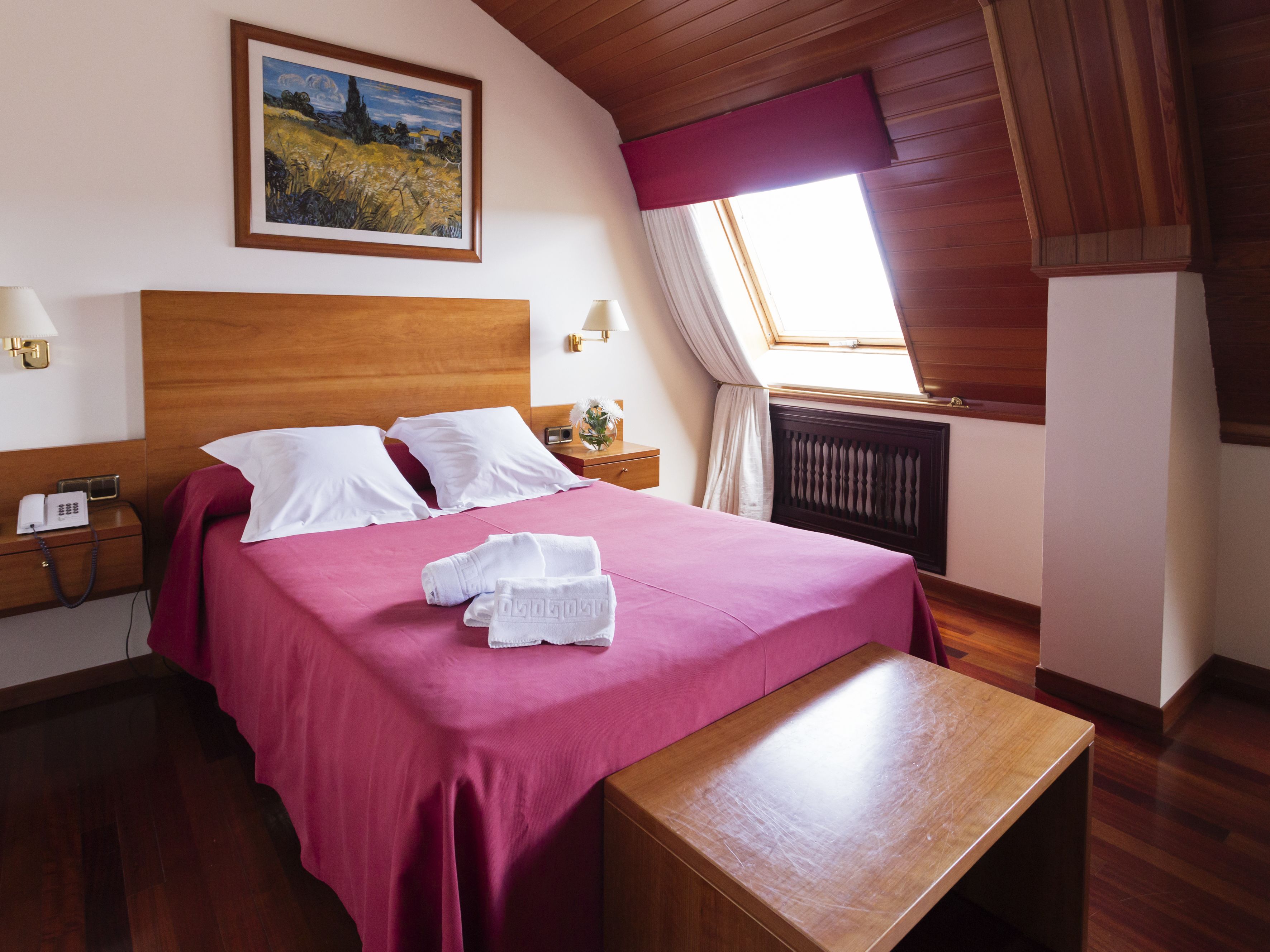 A hotel room with a wooden headboard, a large bed with white linens, a pink bedspread, a wooden nightstand with a lamp, a painting on the wall, and a window with a view of the countryside.