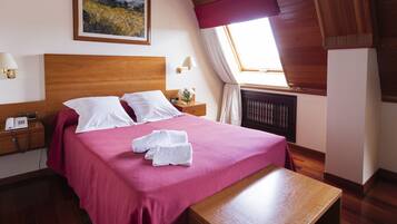 A hotel room with a wooden headboard, a large bed with white linens, a pink bedspread, a wooden nightstand with a lamp, a painting on the wall, and a window with a view of the countryside.