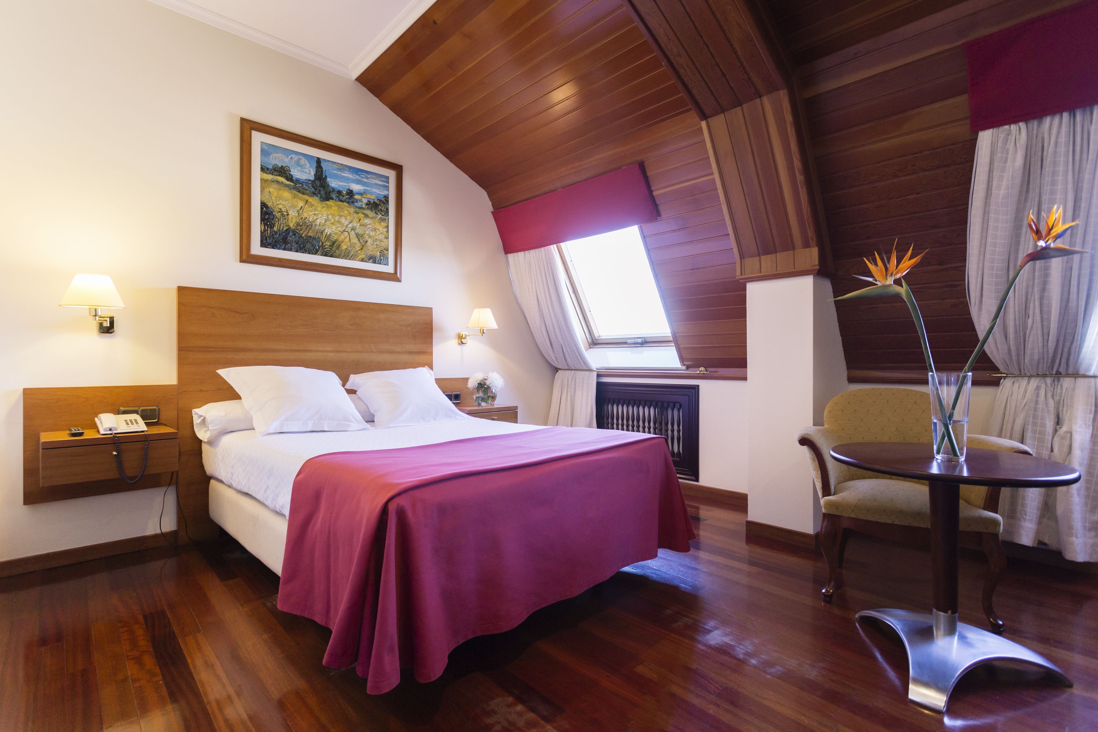 Une chambre d’hôtel avec un plafond en bois, un grand lit recouvert d’une courtepointe rouge, une table de chevet avec une lampe, une chaise et une petite table sur laquelle est posé un vase de fleurs.