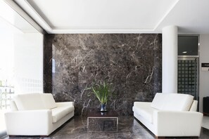 A modern lobby with a marble wall, two white sofas, and a clear glass coffee table with a plant. - Valencia (Ferrol)